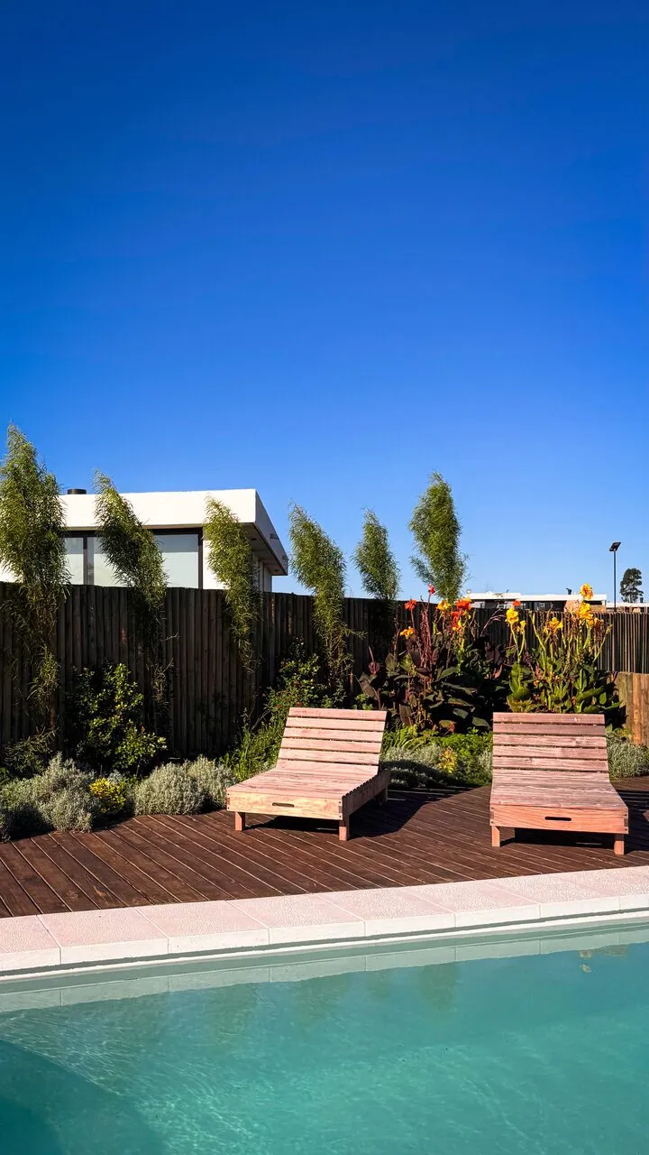 Dos reposeras de madera sobre un deck oscuro junto a la piscina, con achiras rojas y amarillas, plantas plateadas y un cerco de polines